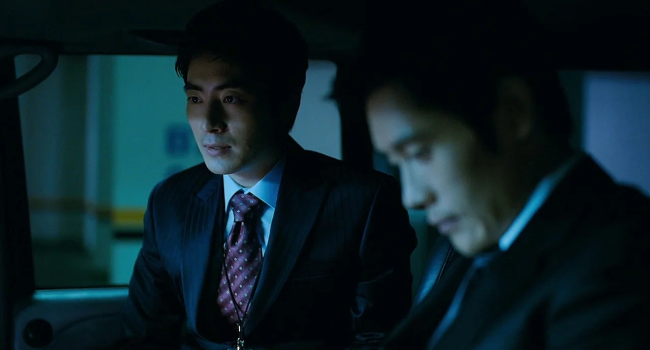 I Saw The Devil (2010) movie still: medium — Two men in business suits sit within the dark, tight interior of a car. One man faces forward while his…