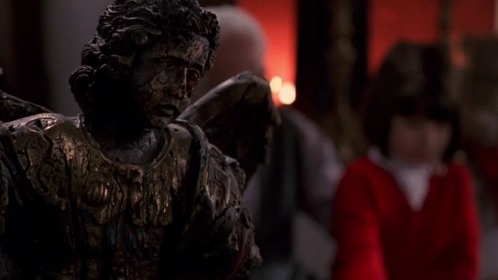 Cronos (1993) movie still: closeup — A dark room features a foreground focus on a weathered wooden angel figurine with damaged features. In the…