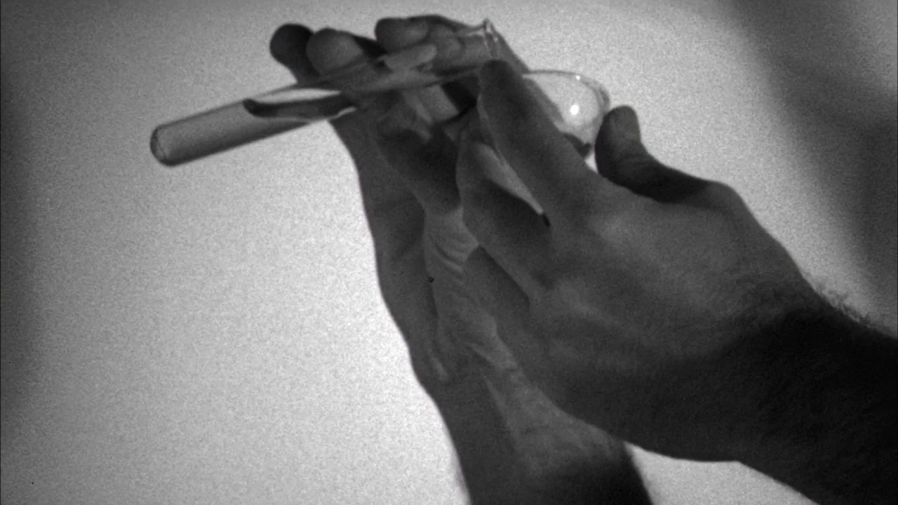 Poison movie still: closeup — A pair of hands manipulates laboratory glassware against a blank, light-colored background. A clear liquid transfers…