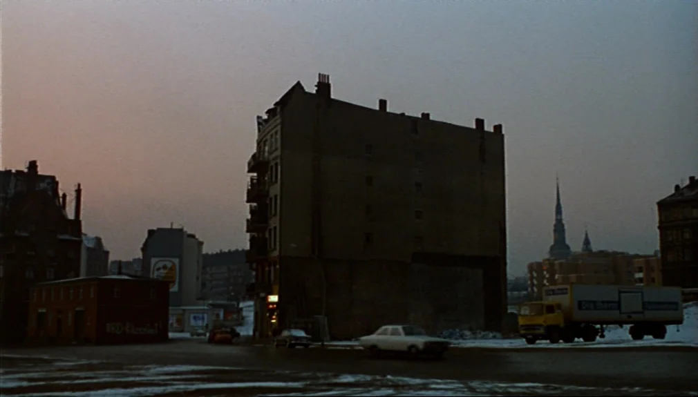 The American Friend (1977) movie still: establishing — A snowy urban intersection features tall apartment blocks and a distant church spire. A…