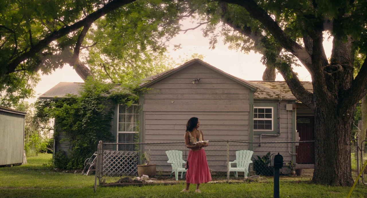 Test Pattern (2019) movie still: fullbody — A grey wood-sided house sits behind a chain-link fence in a grassy yard. A woman stands in the center of…