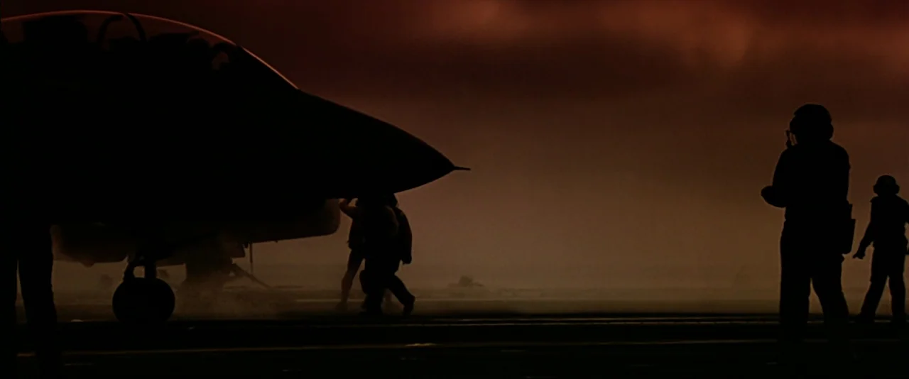 Top Gun (1986) movie still: wide — A dark, atmospheric flight deck at night features the silhouette of a fighter jet. Crew members in uniform move…