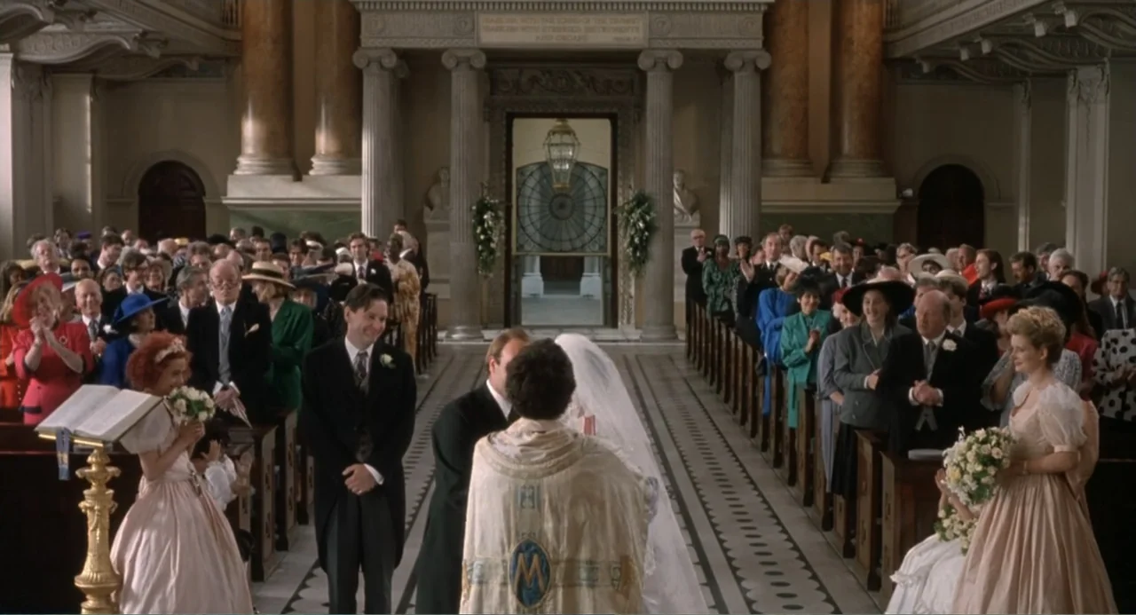 Four Weddings and a Funeral (1994) movie still: fullbody — A stone-columned interior with a central aisle flanked by rows of seated wedding guests.…