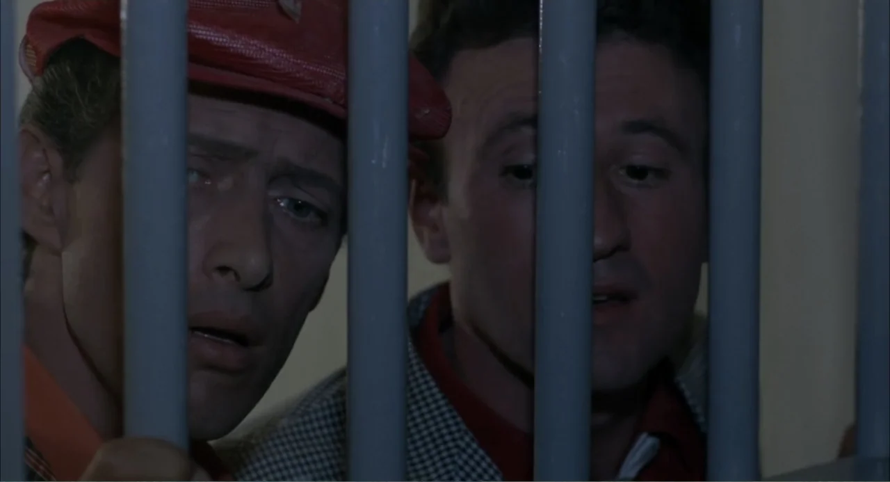 Cottonpickin' Chickenpickers (1967) movie still: medium — A dark, cramped space enclosed by a series of vertical grey iron bars. Two men stand close…