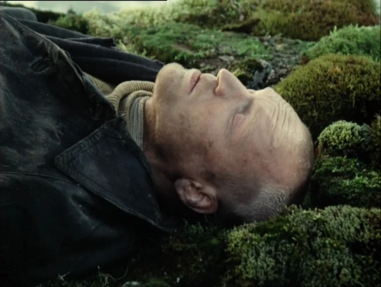 Stalker (1979) movie still: closeup — A man lies on mossy, uneven ground in a tundra environment. The subject is reclined with his eyes closed, his…