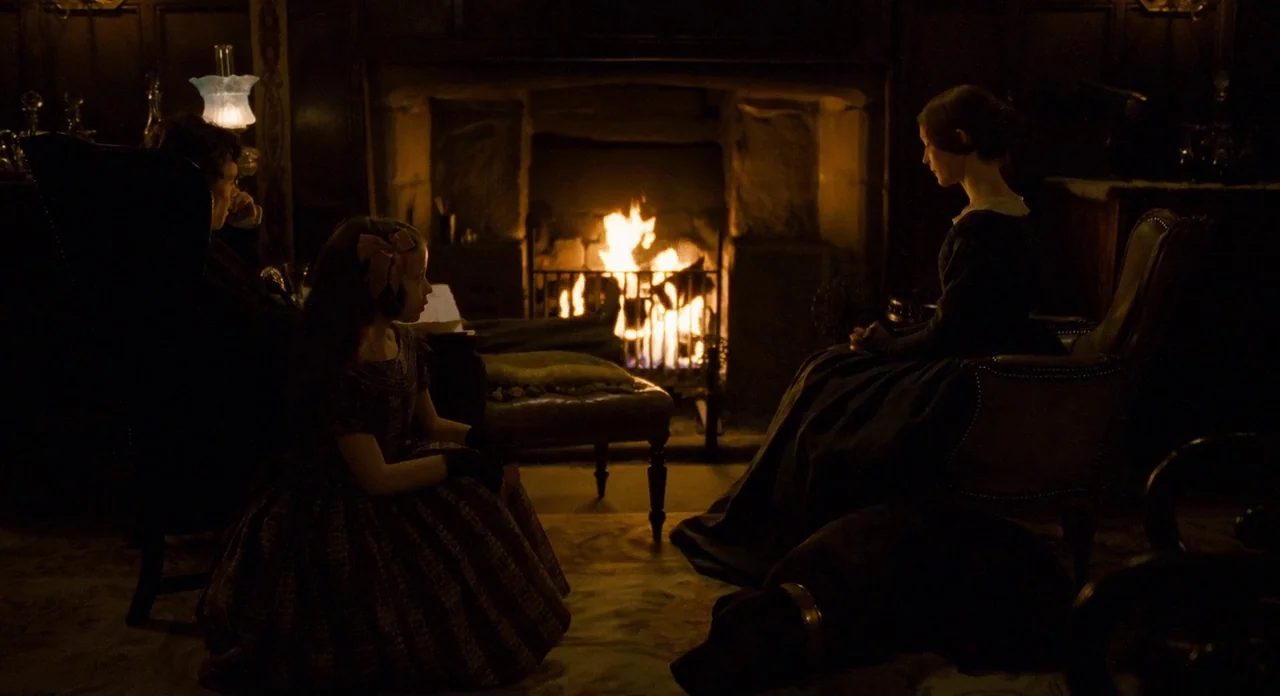 Jane Eyre (2011) movie still: wide — A dark, wood-paneled room features a large stone fireplace with a glowing fire. A woman and a young girl sit…