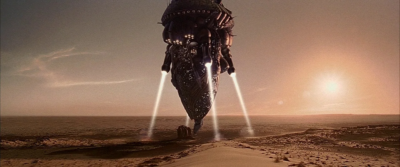 The Fifth Element (1997) movie still: establishing — A vast, empty desert landscape extends toward a low horizon under a glowing sun. An enormous…