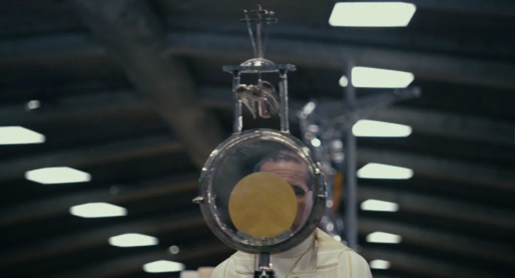 Lourdes movie still: closeup — A man in liturgical vestments holds a gold monstrance inside a large indoor space. The communion host inside the…