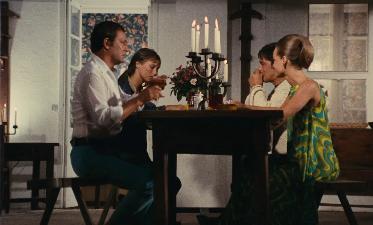 The Swimming Pool (1969) movie still: medium — A rustic interior dining space features a dark wooden table and patterned wallpaper. Four individuals…