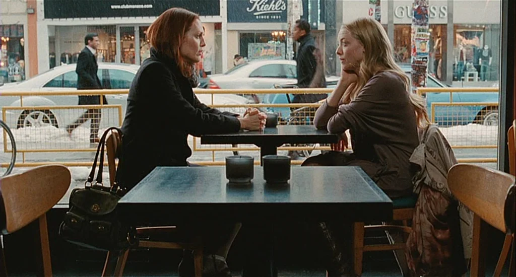 Chloe (2009) movie still: medium — A quiet diner with dark square tables overlooks a snowy city street through windows. Two women sit at a table…