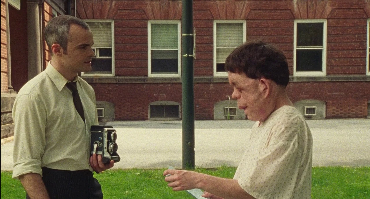 Chained For Life (2018) movie still: medium — An outdoor area features a brick building with multiple windows behind two men. One man holds a…