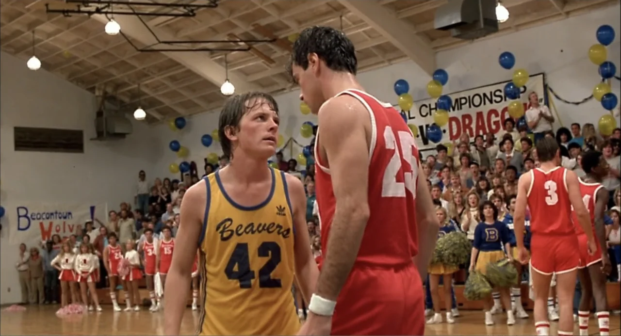 Teen Wolf (1985) movie still: medium — A high school gymnasium decorated with balloons and team banners during a basketball game. Two male players…