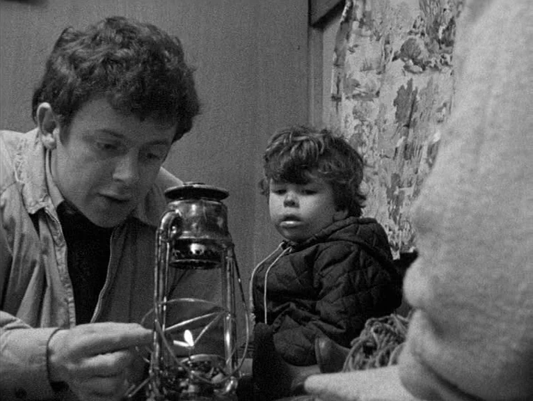 Cathy Come Home (1969) movie still: medium — An interior space features wood-paneled walls and a patterned textile hanging in the background. A man…