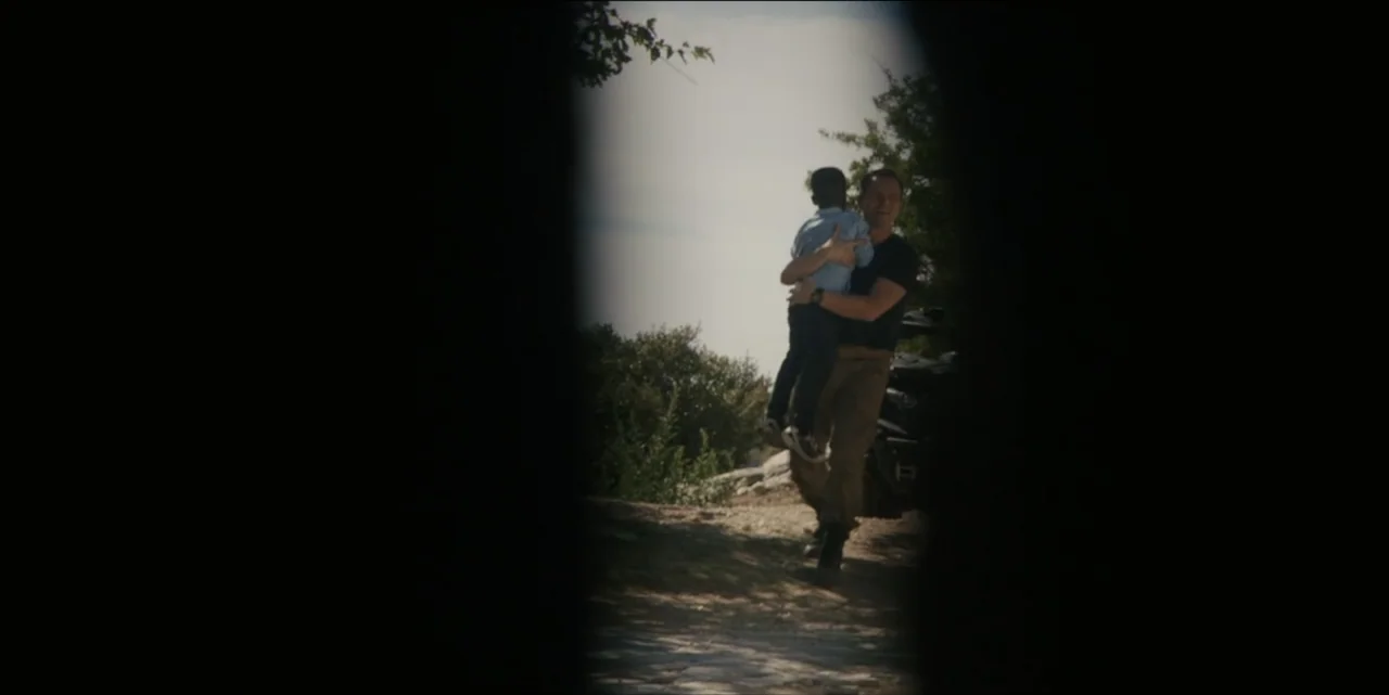 The Hunted (2024) movie still: medium — An outdoor dirt path flanked by dark, out-of-focus foreground elements frame the subjects. An adult male…
