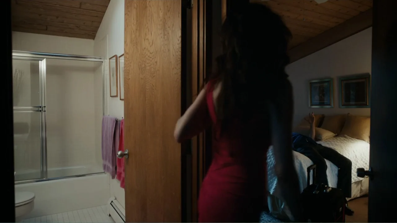 Toss It (2019) movie still: medium — A modest hotel room with wood paneled ceilings connects to a tiled bathroom. A woman in a red dress walks…