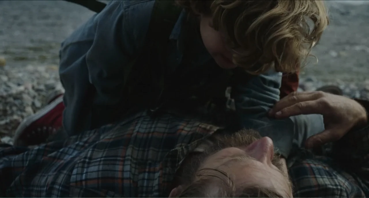 Cowboys (2014) movie still: medium — A rocky shoreline sits beside dark water under overcast sky. A child leans over a bearded man resting on the…