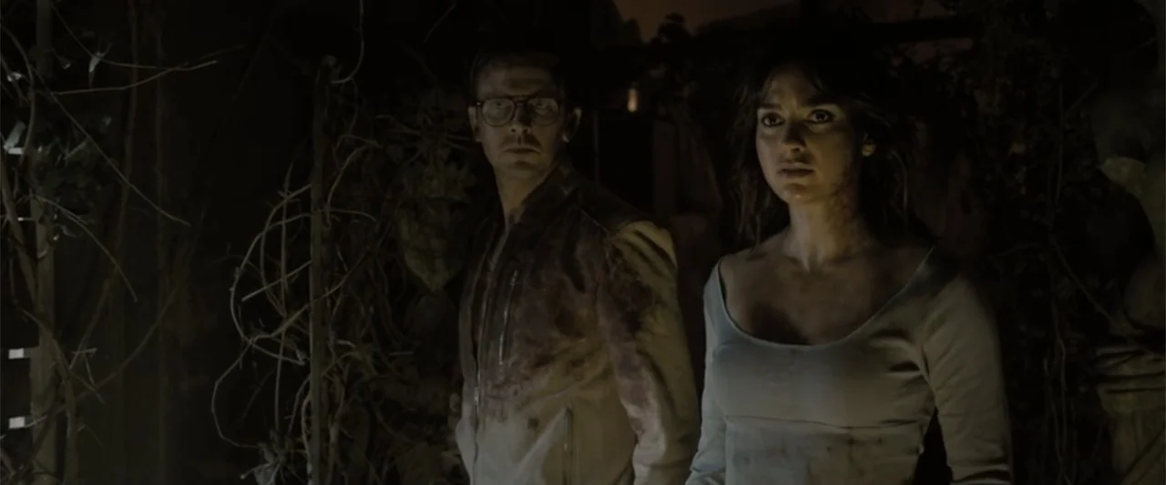 Abigail (2024) movie still: medium — Two individuals stand in a dark, foliage-covered space with indistinct wooden structural elements behind them.…