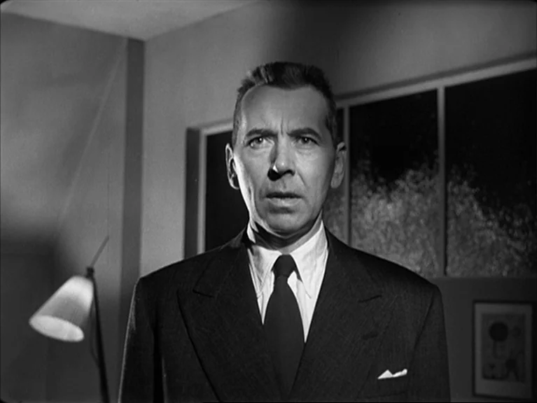 The Sleeping Tiger (1954) movie still: medium — A dark study featuring a floor lamp and framed wall art. A man in a suit stands in the center of the…