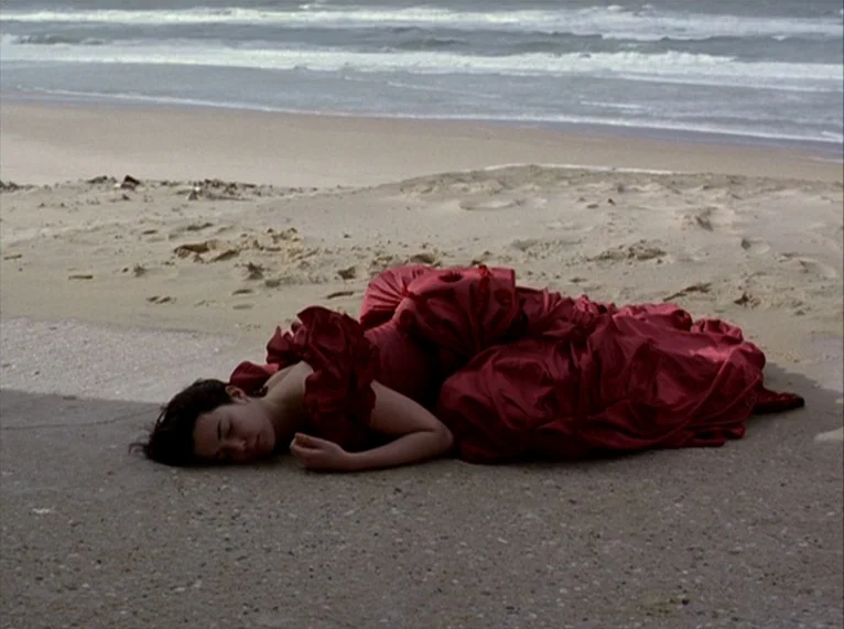 For Ever Mozart (1996) movie still: fullbody — An expanse of wet sandy beach borders the breaking waves of an ocean. A woman wearing a formal red…
