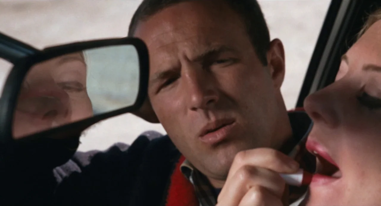 The Rain People (1969) movie still: closeup — The interior of a car shows a woman applying lipstick in the rearview mirror. A man sits beside her…