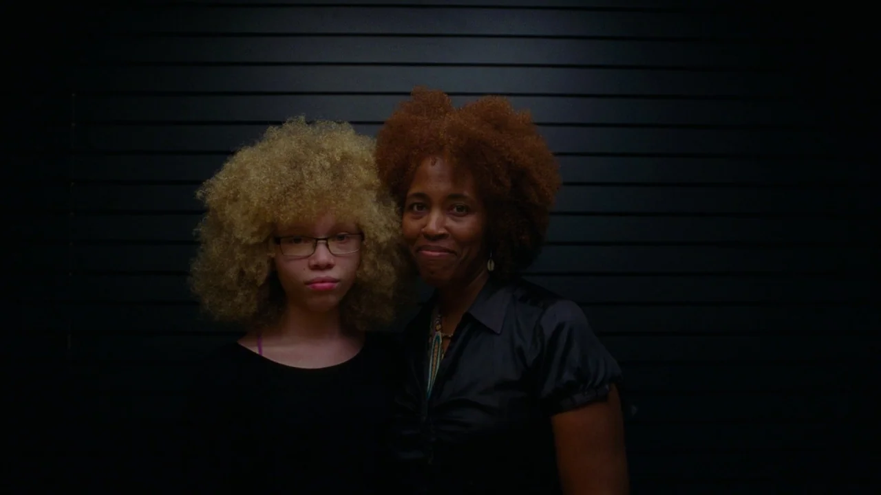 Pass Over (2018) movie still: medium — A portrait of two women stands before a dark, horizontal-slatted wall in a shop. They pose together while…