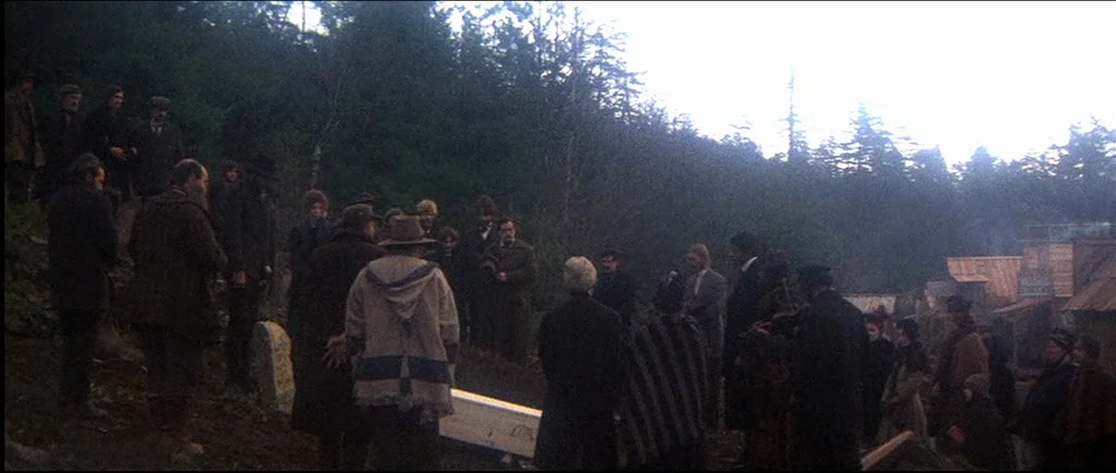 McCabe & Mrs.Miller (1971) movie still: wide — A group of people stands in a clearing within a dense, pine-covered mountain forest. They are…