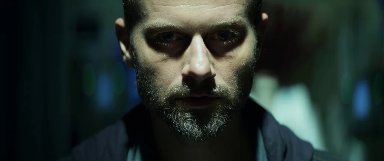 The Empty Man (2020) movie still: closeup — A dark, dimly lit corridor background is heavily out of focus. A man with a beard stares directly…