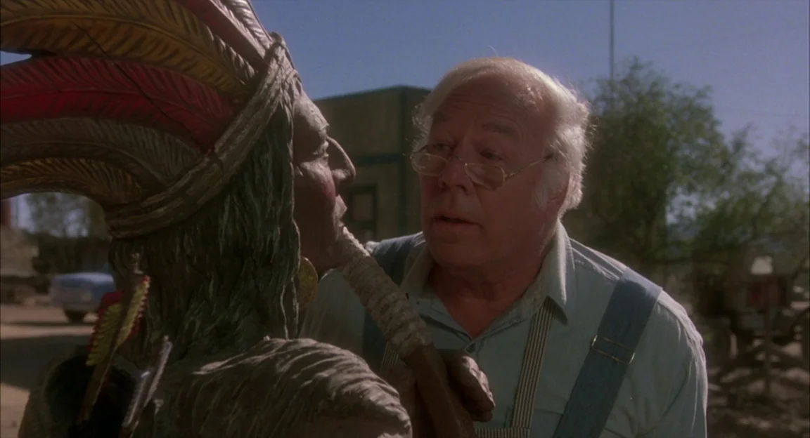 Creepshow 2 (1987) movie still: medium — An elderly man stands in a dusty desert town square beside a statue. He holds onto the wooden statue while…