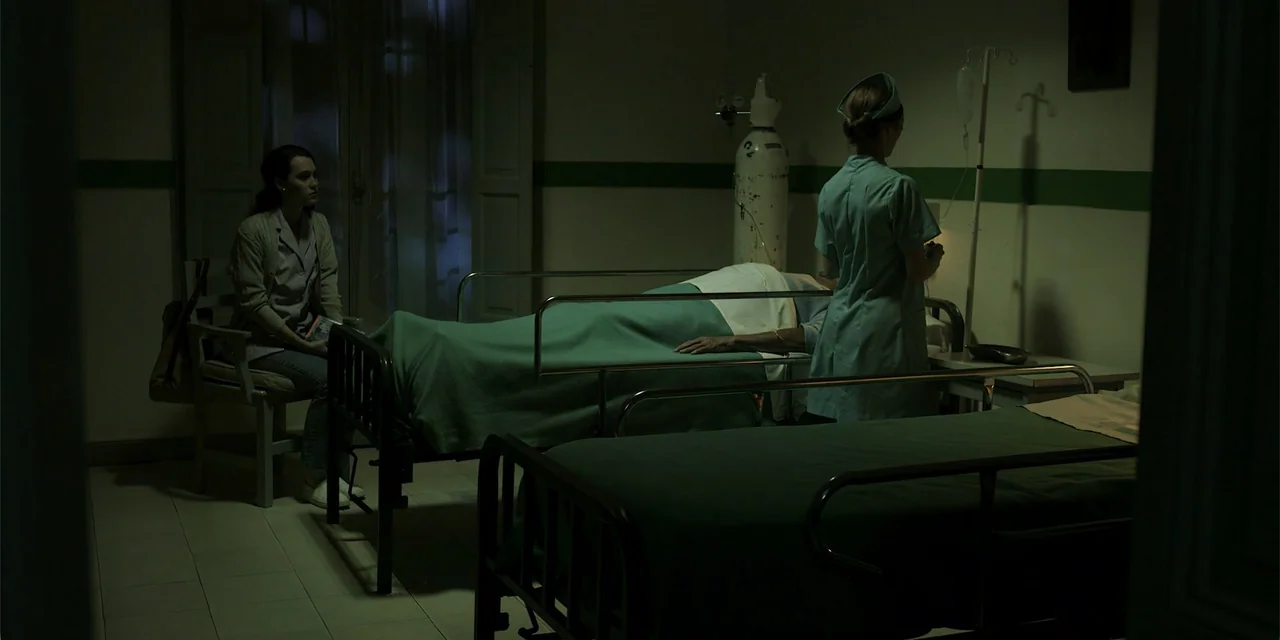 La coda del diavolo (2024) movie still: wide — A dimly lit hospital room contains two metal bed frames and an oxygen tank. A visitor sits quietly…