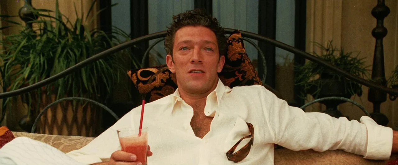 Ocean’s Twelve (2004) movie still: medium — A man sits on an outdoor patio bench with a wrought iron backrest and foliage. He holds a glass with a…