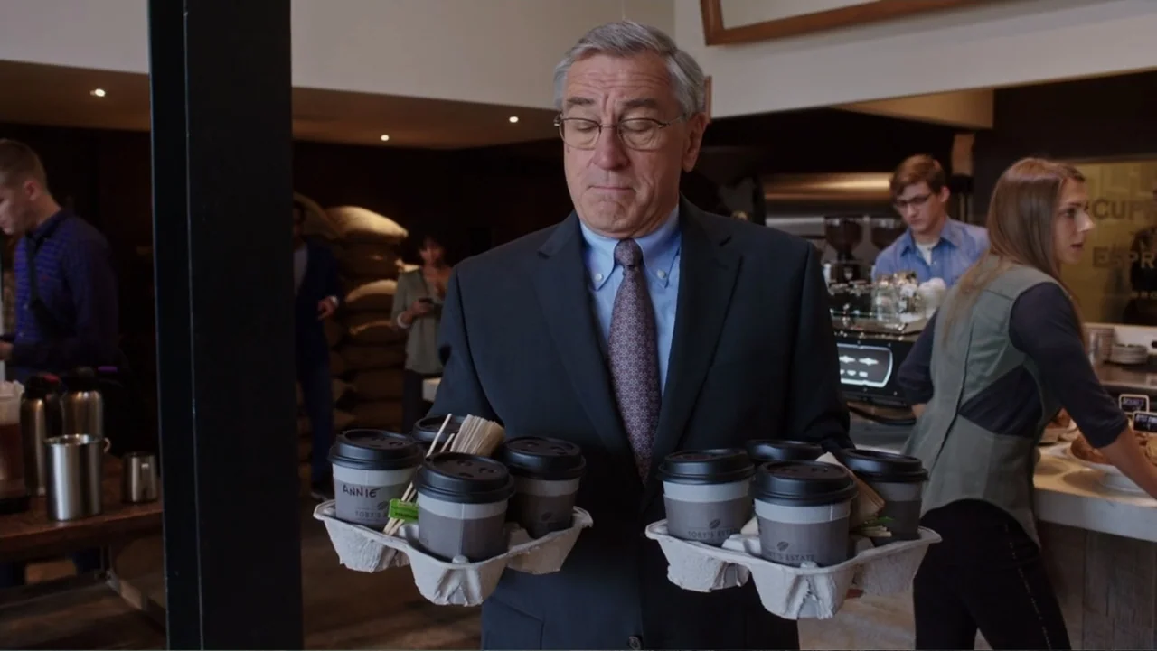 The Intern (2015) movie still: medium — A bustling coffee shop interior with other customers visible in the blurred background. A mature man in a…