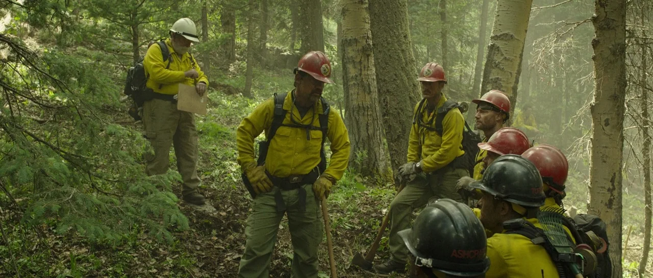 Only The Brave (2017) movie still: fullbody — A group of firefighters wearing yellow shirts and hard hats stands in a forest. They appear to be…