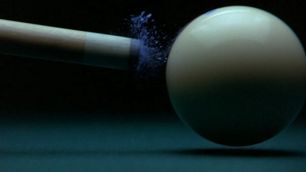 The Colour of Money (1986) movie still: closeup — A pool cue tip makes contact with the surface of a white billiard cue ball. Particles of blue…