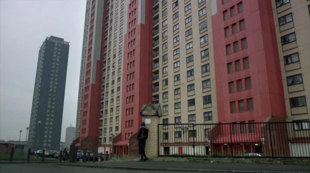 Red Road (2006) movie still: wide — A towering high-rise apartment block with red accents dominates the urban landscape. A person stands alone…