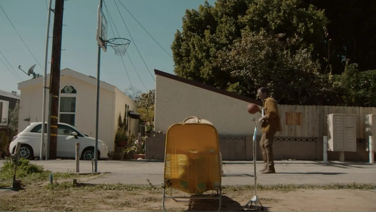 The Cleaner (2021) movie still: fullbody — A sunny residential lot in a mobile home park features a basketball hoop. An older man plays with a…