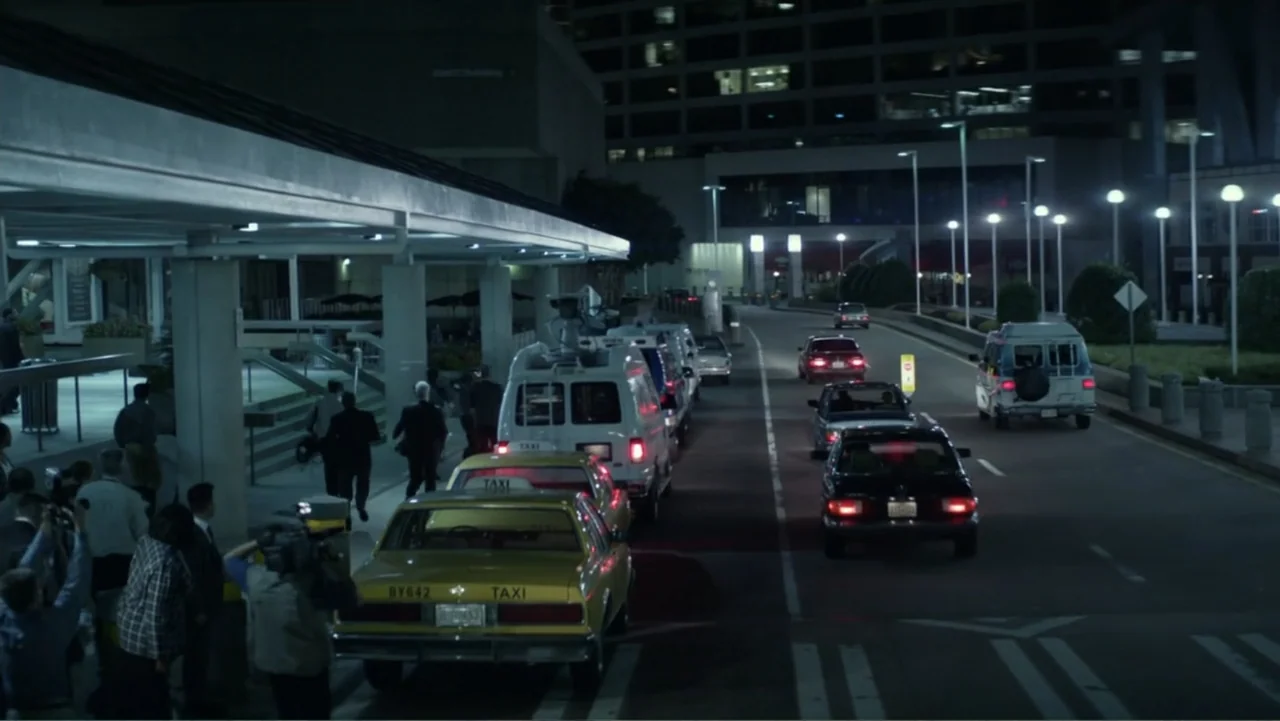 Confirmation (2016) movie still: wide — An airport terminal exterior at night features a paved driveway lined with taxis and vehicles. A small group…