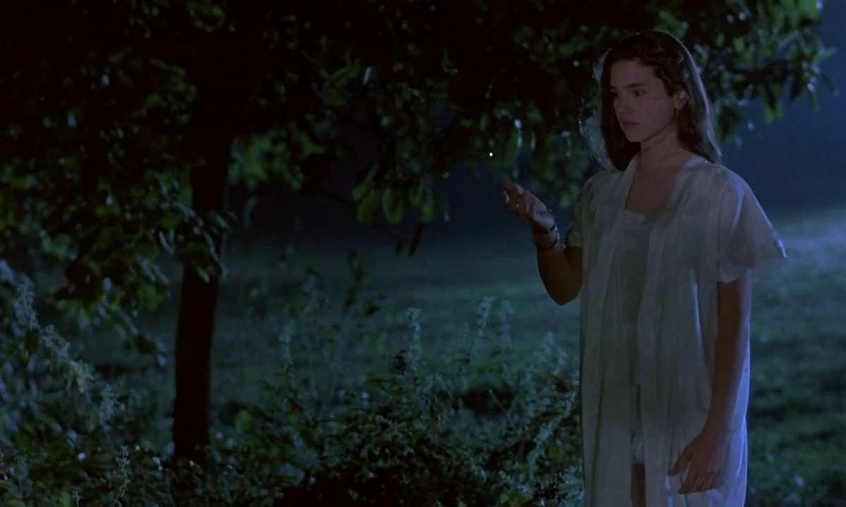 Phenomena (1985) movie still: medium — A dark, moonlit outdoor area contains a single tree and low dense shrubbery. A young woman in a white…