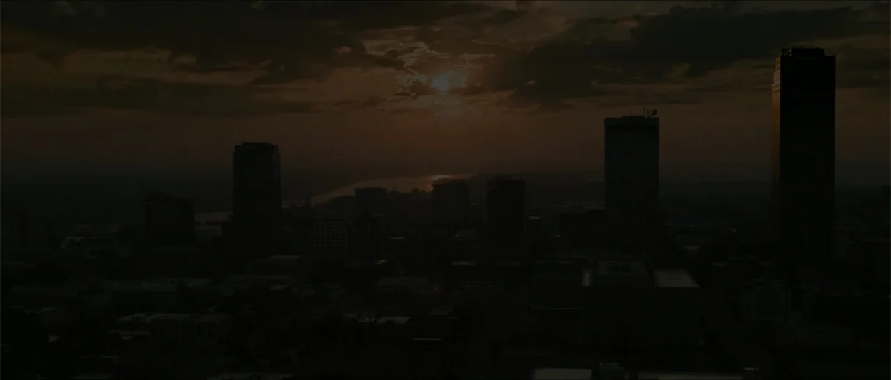 Crescent City (2024) movie still: establishing — A dark city skyline is silhouetted against a cloudy sunset over a distant river. The sun is…