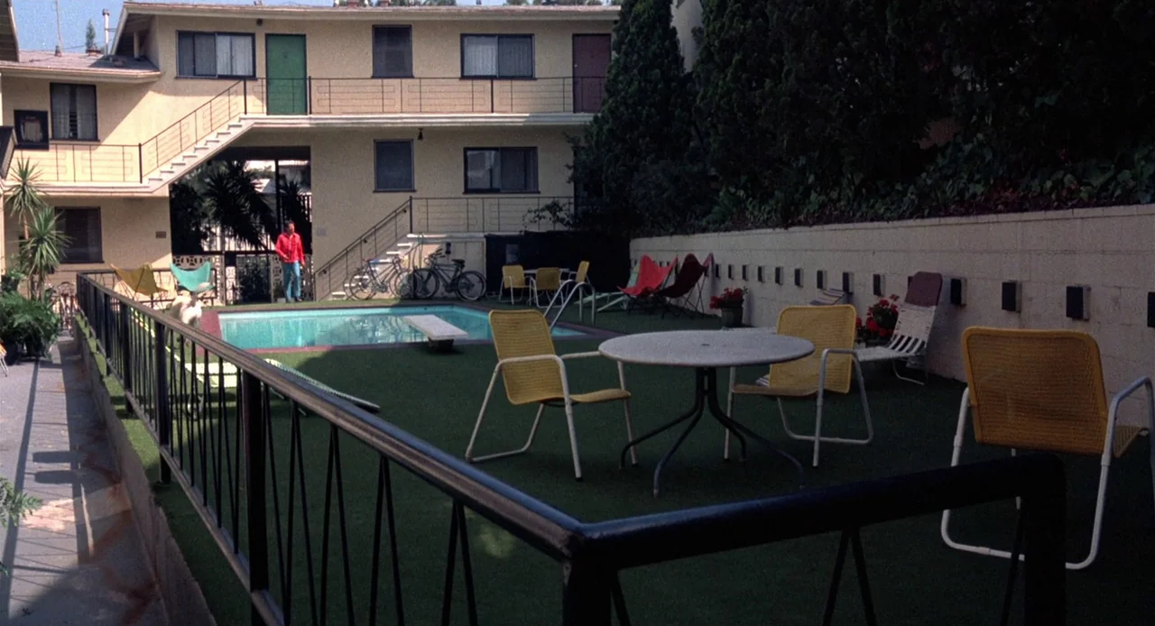 Breathless (1983) movie still: wide — A suburban apartment complex features a small pool area with green outdoor carpeting. A lone man in a red…