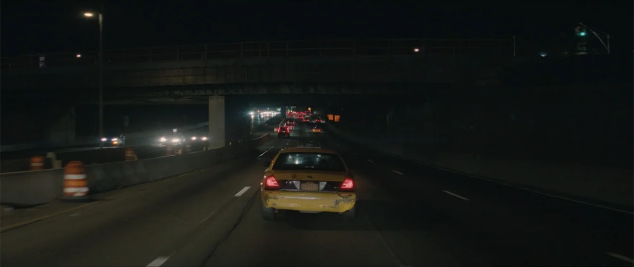 Daddio (2024) movie still: wide — A yellow taxi drives on a multi-lane highway underneath a large concrete bridge overpass. The road features orange…
