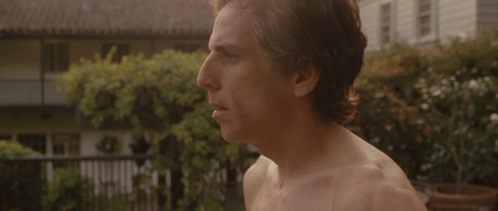 Greenberg (2010) movie still: medium — A residential courtyard features a two-story building in the background with greenery. A man stands shirtless…