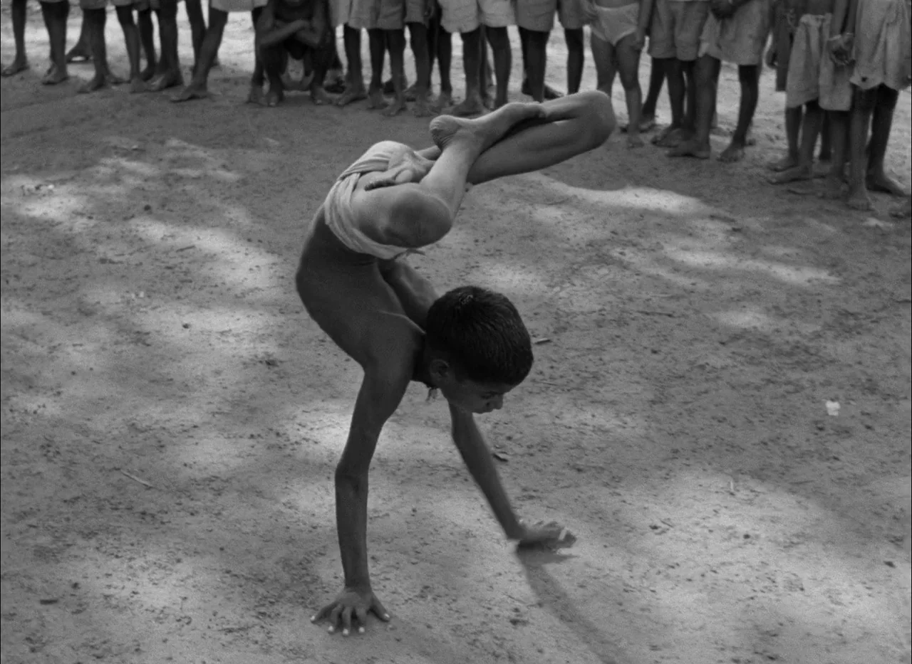 Aparajito (1956) movie still: fullbody — An open dirt lot filled with a crowd of standing children encircling a performer. A young boy in a…