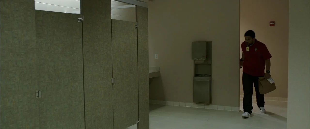 Den of Thieves (2018) movie still: medium — A public restroom with stall partitions and a wall-mounted paper towel dispenser. A man in a red polo…
