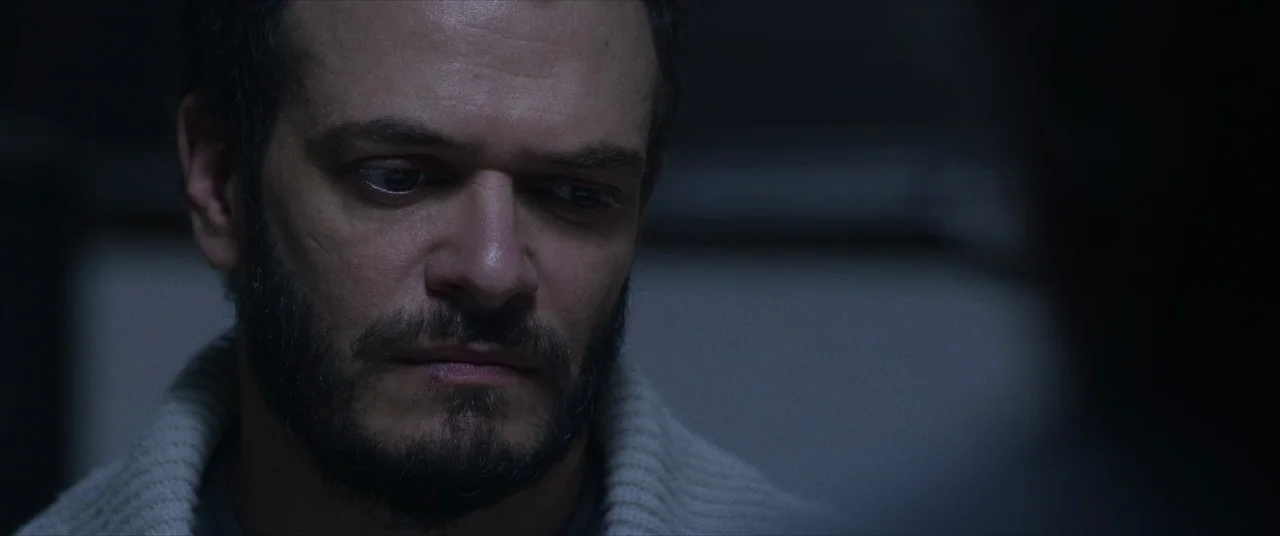 Terrified (2017) movie still: closeup — A dimly lit interior space with a dark, indistinct background. A man wears a ribbed sweater and gazes down…