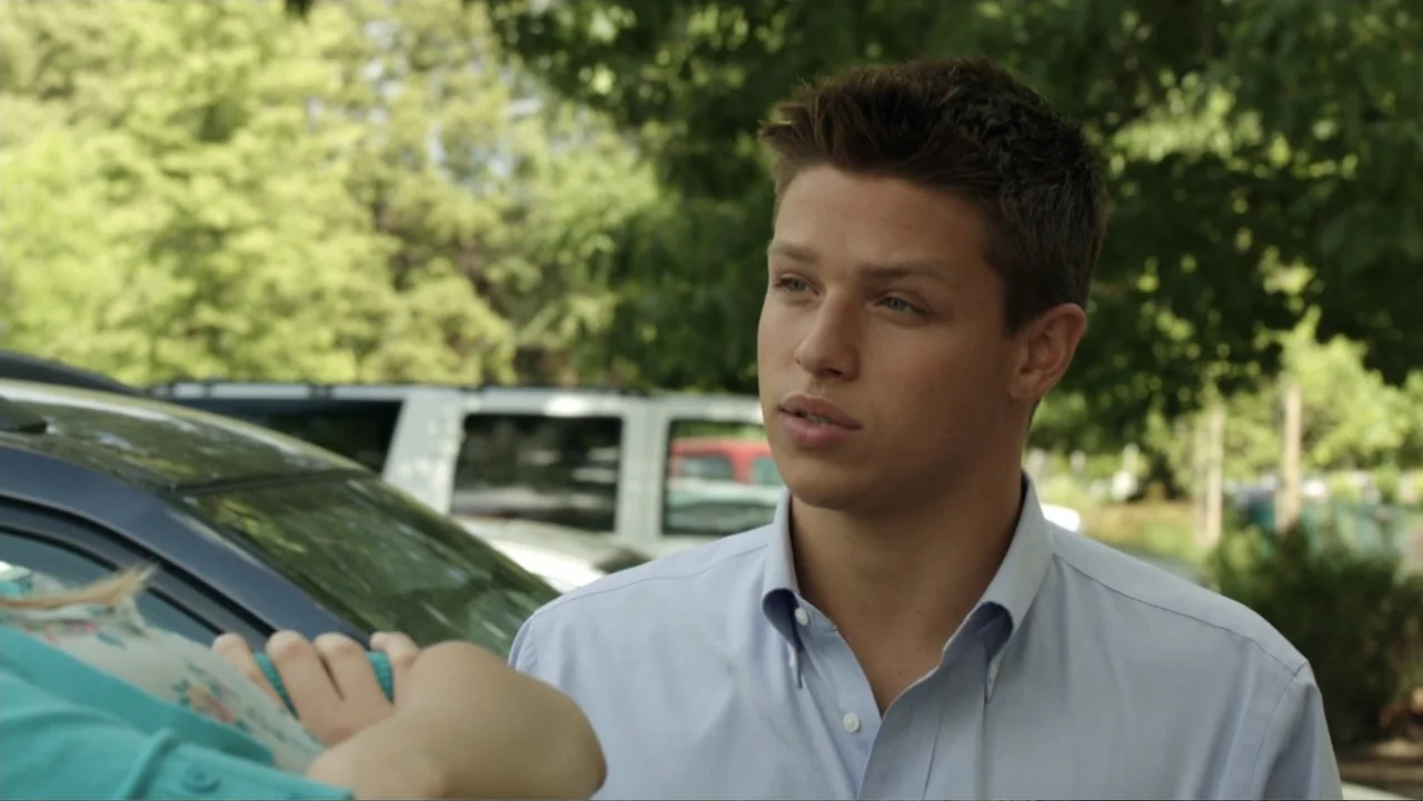 At Middleton (2013) movie still: medium — A sunny outdoor parking lot with green trees visible in the soft-focus background. A young man wearing a…