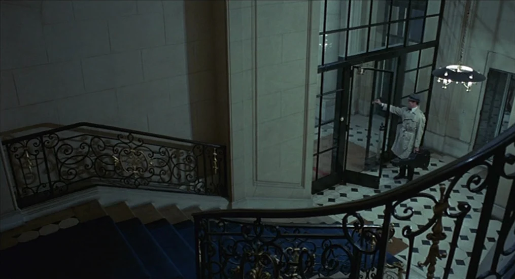 Le Cercle Rouge (1970) movie still: wide — An expansive marble-floored lobby features a grand staircase with ornate metal railings. A man in a…
