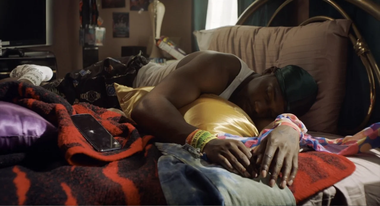 House Party (1990) movie still: medium — A bedroom contains a bed with a large metal headboard and colorful patterned blankets. A man wearing a…