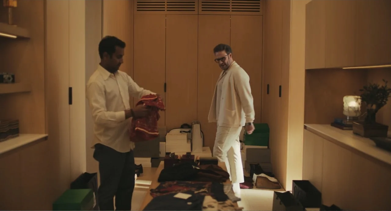 Good Fortune (2025) movie still: medium — A spacious walk-in closet with light wood paneling and stacked storage boxes. Two men stand facing each…