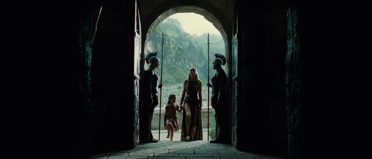 Wonder Woman (2017) movie still: wide — A stone arched doorway frames a view of a lush mountain landscape outside. A woman and child walk through…