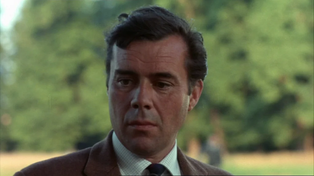 Accident (1967) movie still: closeup — A man stands outdoors in front of a blurry background of trees. The subject looks toward the left side of the…